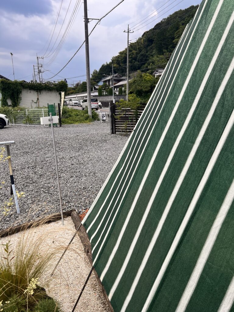 A photo of the store taken from the Little Valley parking lot. Part of the green striped sunshade is visible, and a lush landscape spreads out in the background. You can see an unpaved gravel parking lot.