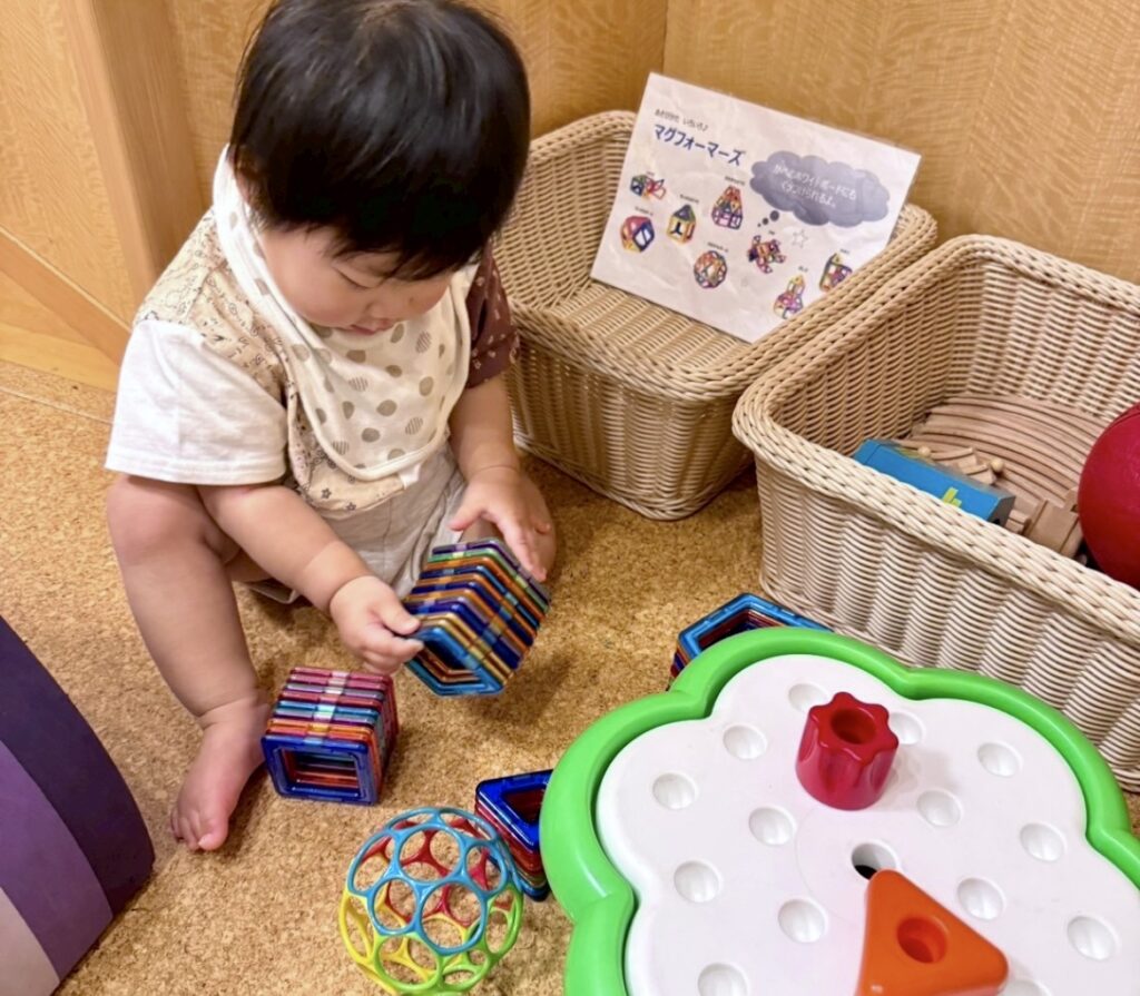 マグフォーマーなどの知育おもちゃで遊ぶ赤ちゃん。おもちゃ収納用のカゴが背景に見える。