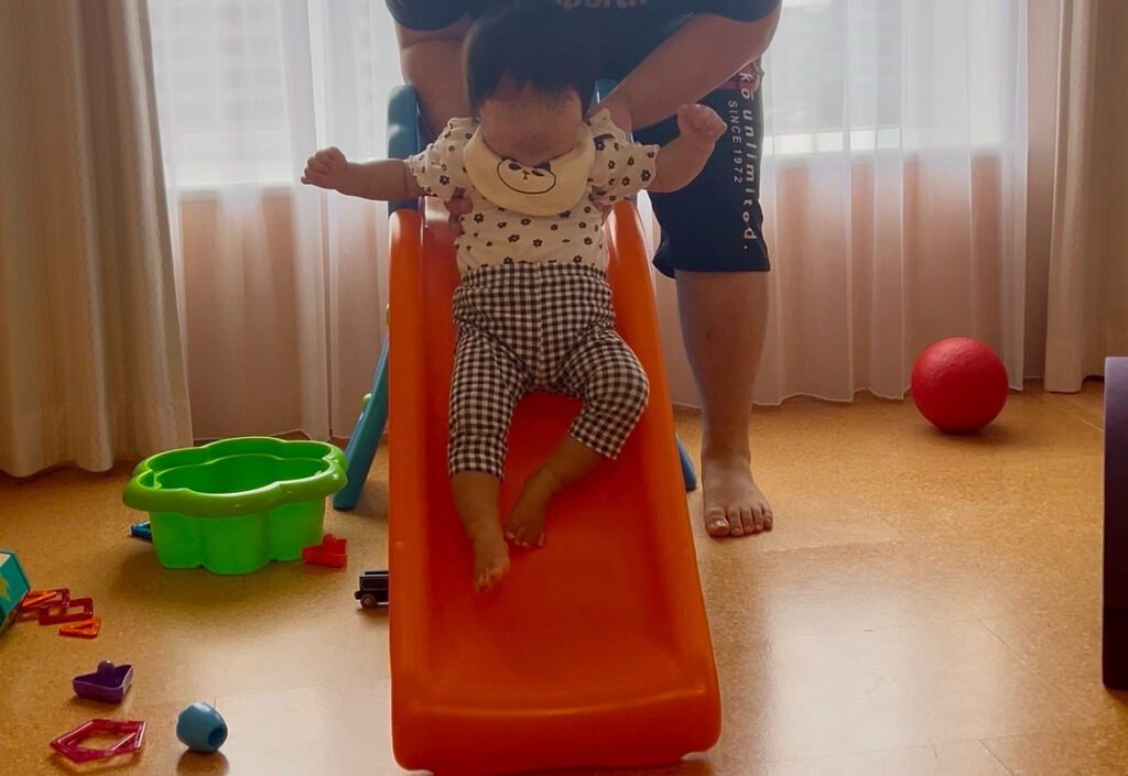  室内の滑り台で遊ぶ赤ちゃん。保護者が後ろからサポートしている様子。